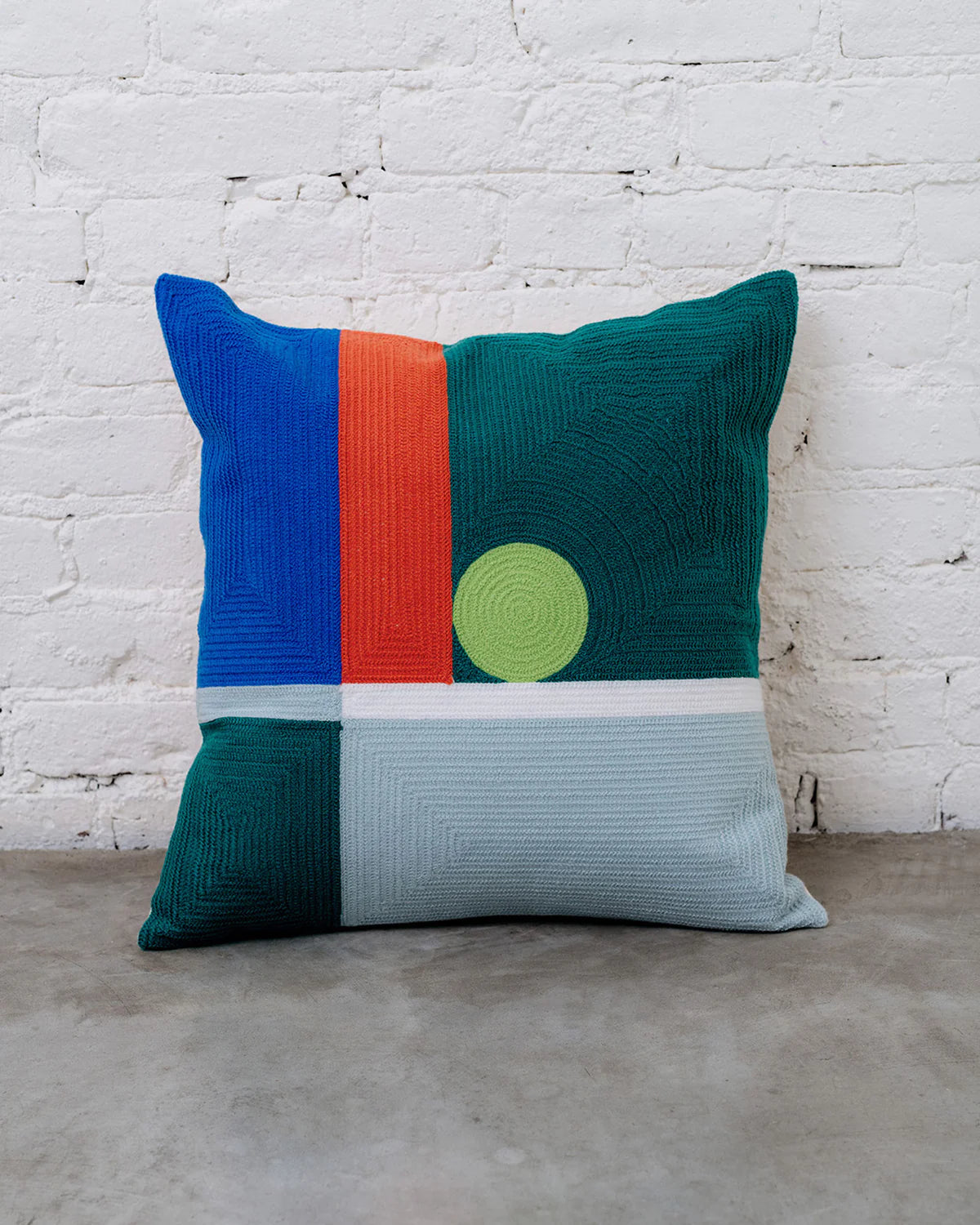 coussin dusendusen design moderne graphique verte foncé, vert clair, bleu et rouge posé par terre sur fond de mur en briques blanches