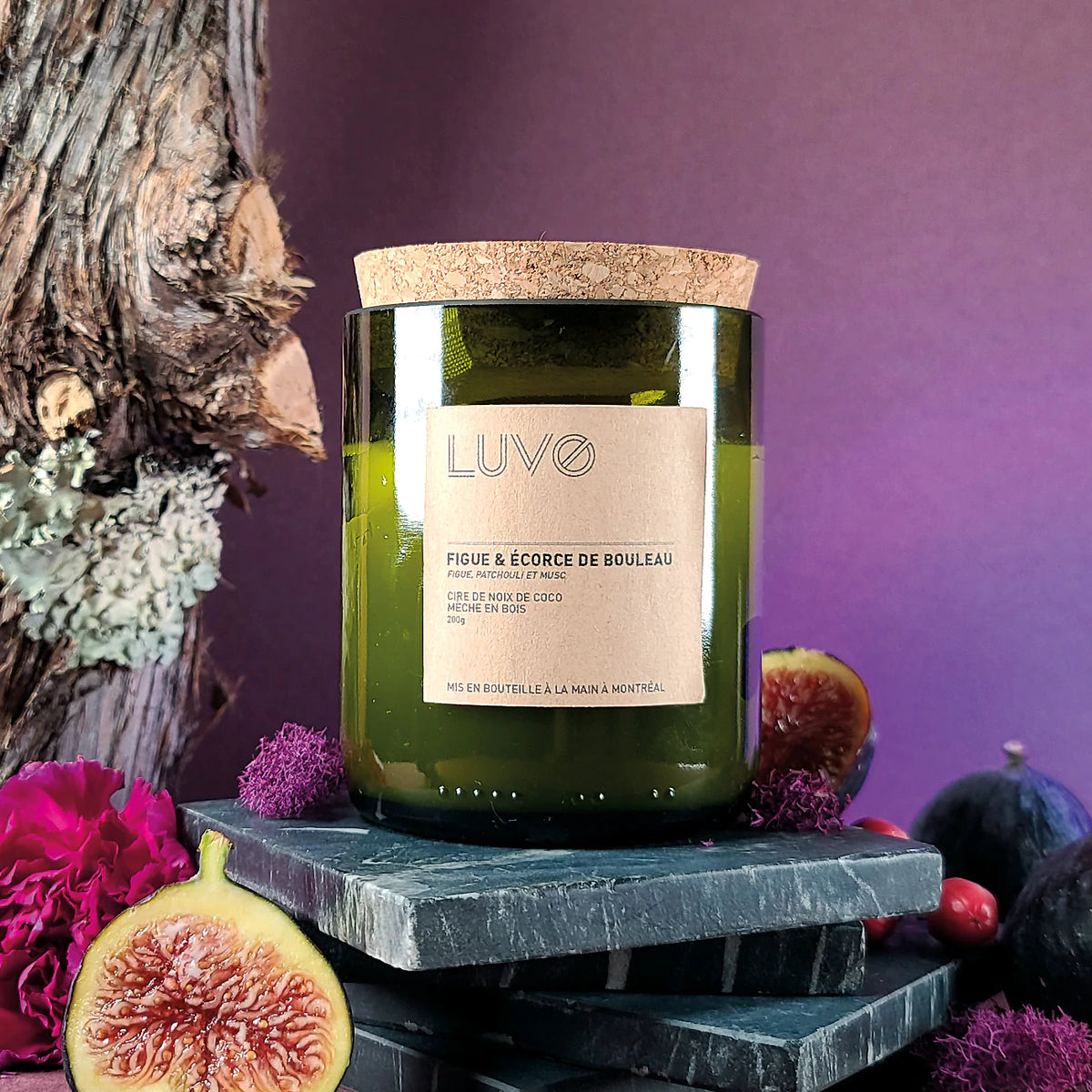 bougie Luvo parfumée figue et écorce de bouleau. Bougie dans un bocal de bouteille vert de vin et bouchon en liège sur fond violet avec un arbre et des figues