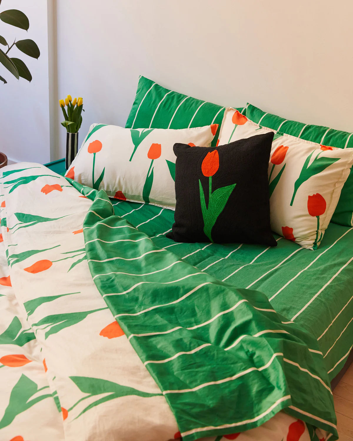 coussin noir avec une tulipe rouge et verte brodé. Le coussin est posé sur un lit avec des draps rayés verts et blancs et d'autres coussins tulipe blanc rouge et vert