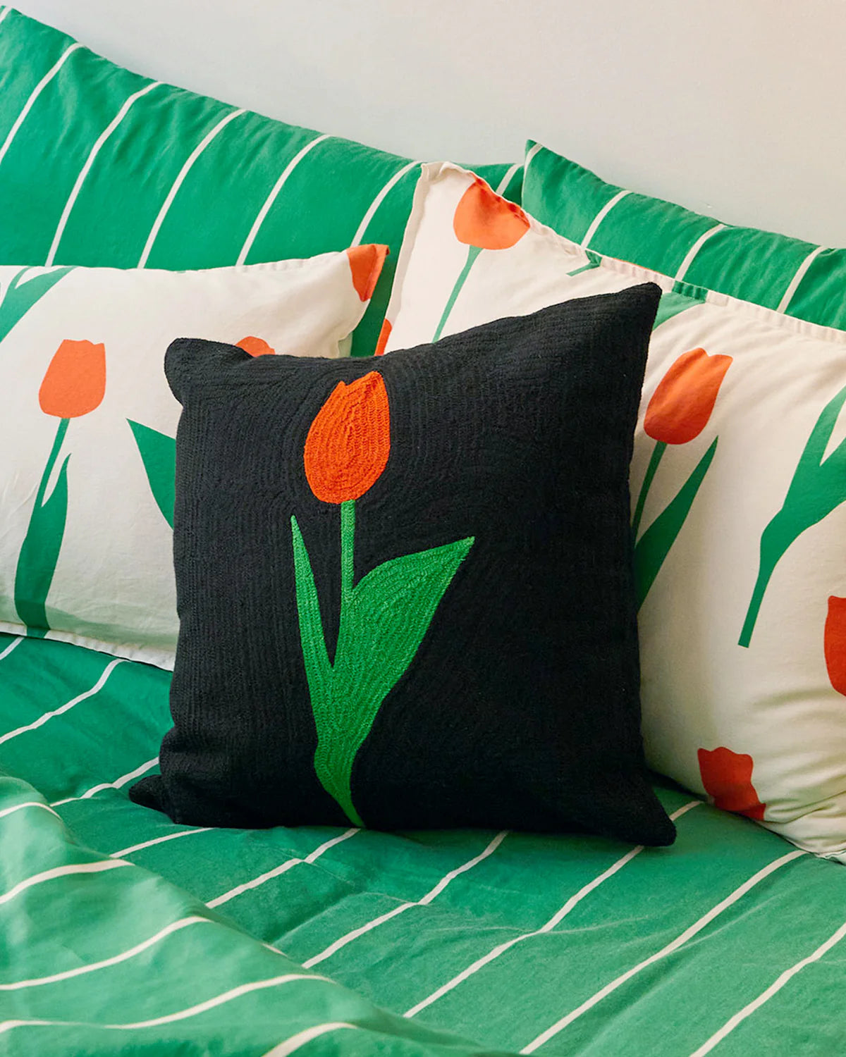 coussin noir avec une tulipe rouge et verte brodé. Le coussin est posé sur un lit avec des draps rayés verts et blancs et d'autres coussins tulipe blanc rouge et vert